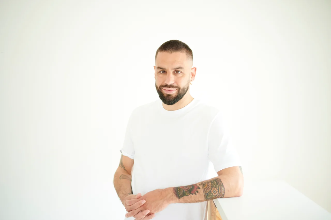 Antoine Andrieu, en t-shirt blanc, regard caméra et tatouages visibles.